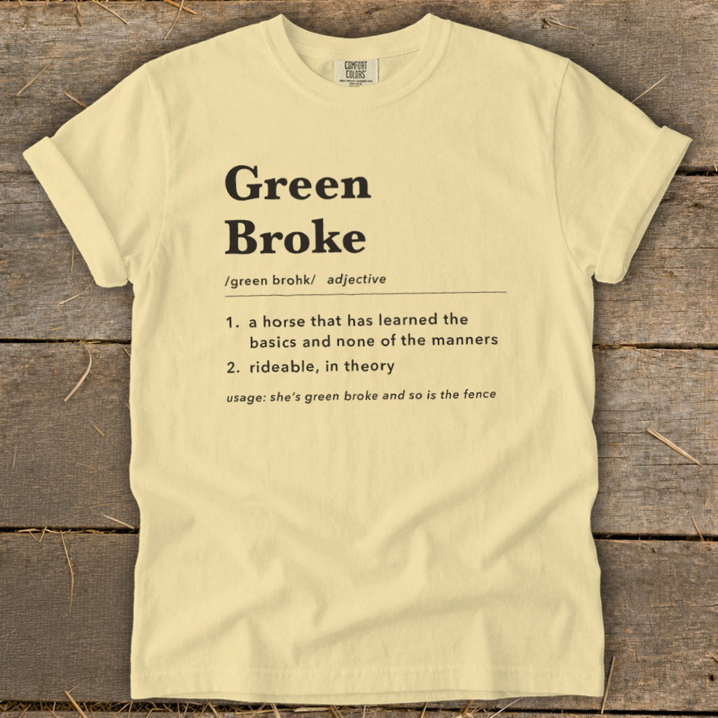 Light yellow t-shirt laid flat on wooden planks, featuring black text defining "Green Broke" as an adjective.