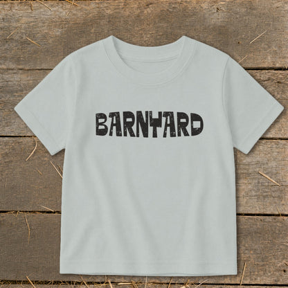 A light gray short-sleeved child's t-shirt lies flat on a rustic wooden surface, featuring the word BARNYARD printed in distressed black letters.