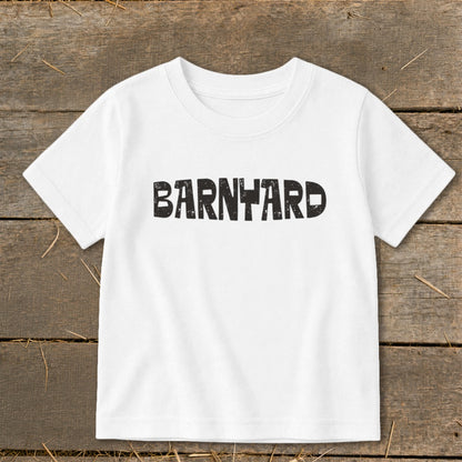 White children's short-sleeve t-shirt with black distressed lettering spelling "BARNYARD" centered on the chest, laid flat on a rustic wooden background.