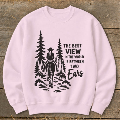 A light pink crewneck sweatshirt features a black graphic of a horse rider in the woods with text reading The Best View In The World Is Between Two Ears.