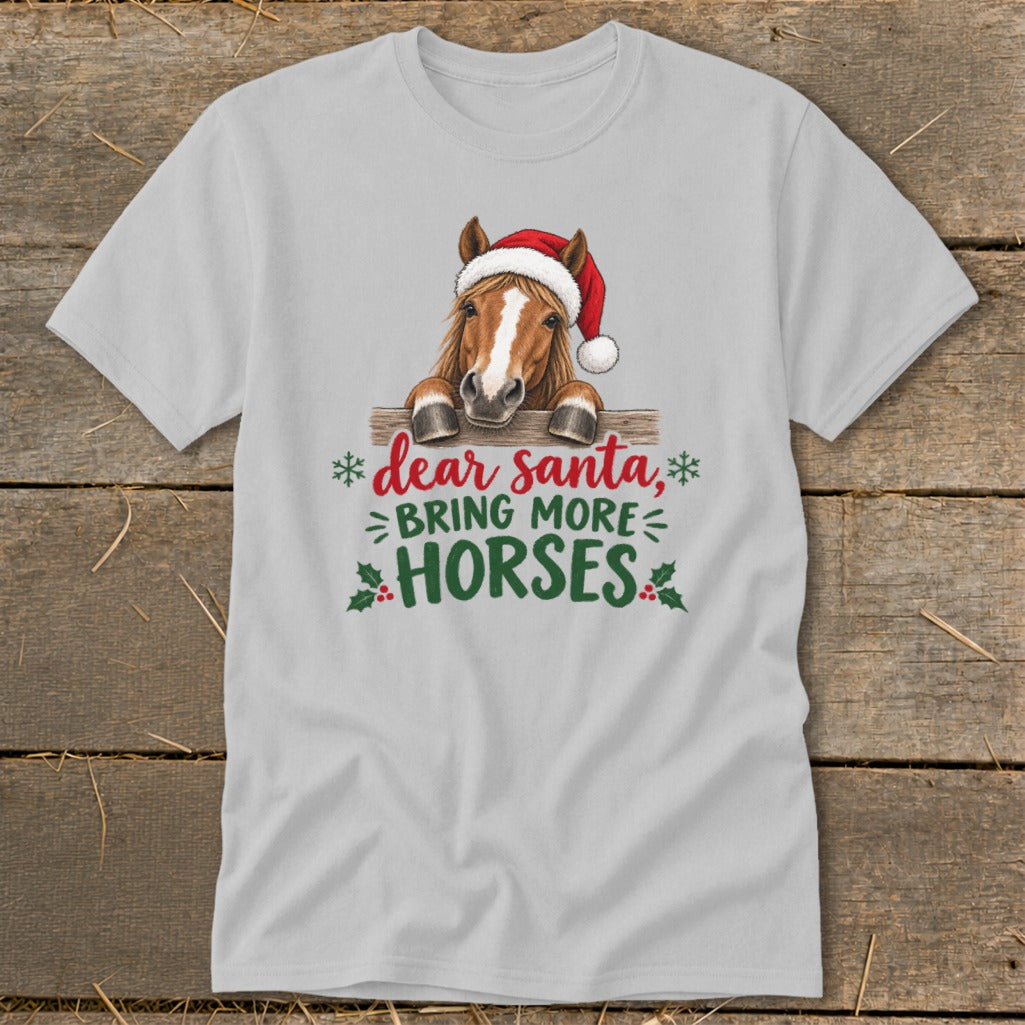 A light gray t-shirt featuring a graphic of a horse in a Santa hat behind a wooden fence with text reading Dear Santa, Bring More Horses.
