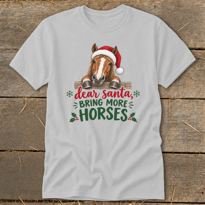 A light gray t-shirt featuring a graphic of a horse in a Santa hat behind a wooden fence with text reading Dear Santa, Bring More Horses.
