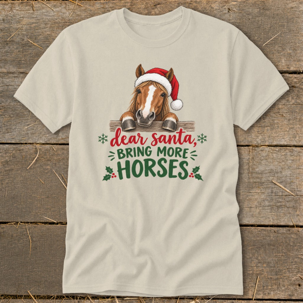 A cream-colored t-shirt with a graphic of a horse wearing a Santa hat behind a fence, above the text Dear Santa, Bring More Horses.