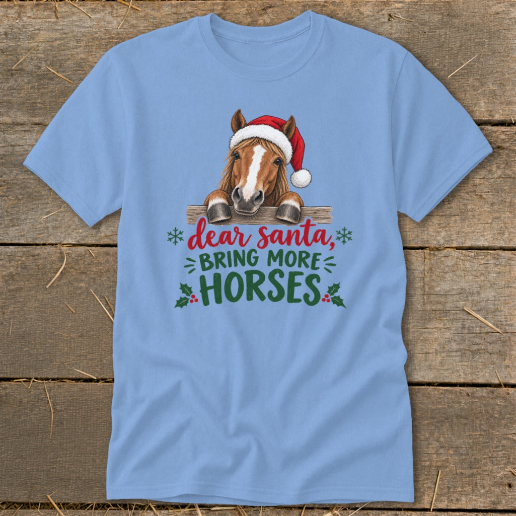 A light blue t-shirt featuring a horse wearing a Santa hat behind a wooden fence with text saying, "Dear Santa, Bring More Horses."