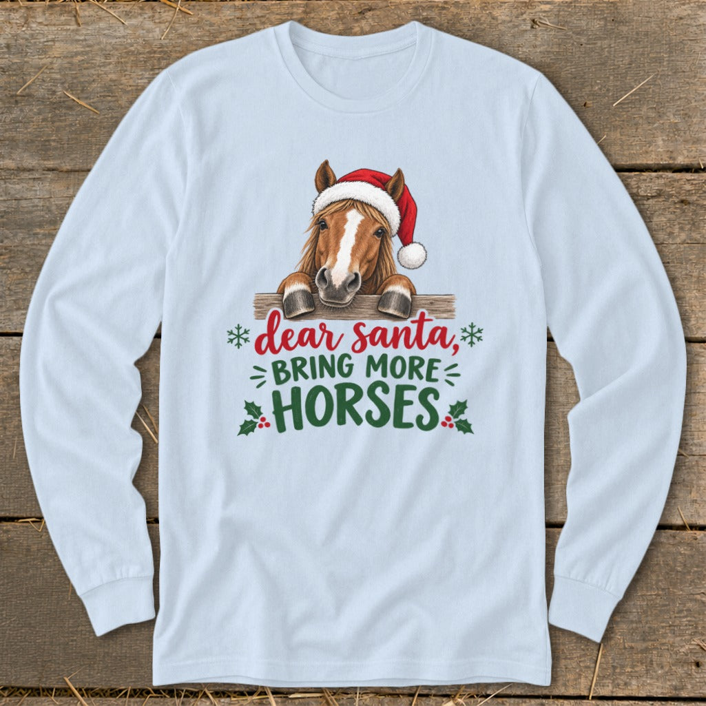 A light blue long-sleeve t-shirt featuring an illustration of a horse wearing a Santa hat behind a wooden fence with the text "dear santa, bring more horses".