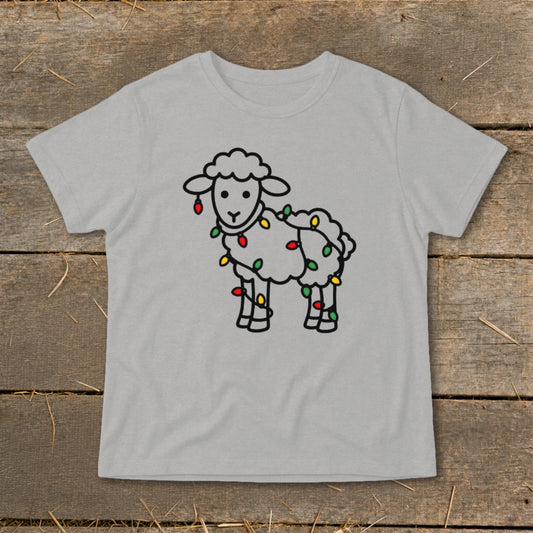 A light gray t-shirt featuring a black outline drawing of a fluffy sheep wrapped in colorful Christmas lights, laid flat on a rustic wooden surface.