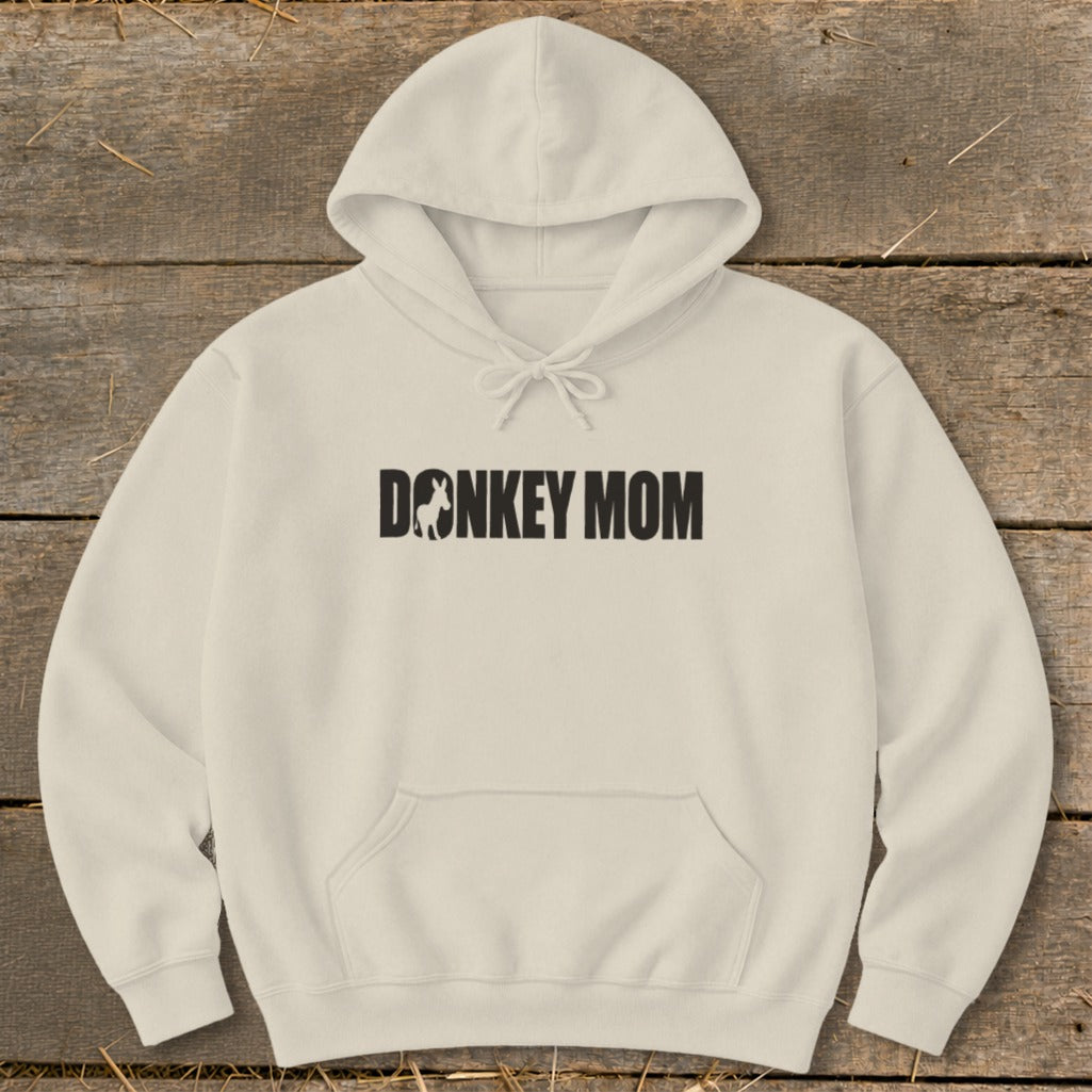 A cream-colored hooded sweatshirt featuring the text DONKEY MOM with a donkey silhouette inside the letter O, laid flat on a wooden surface.