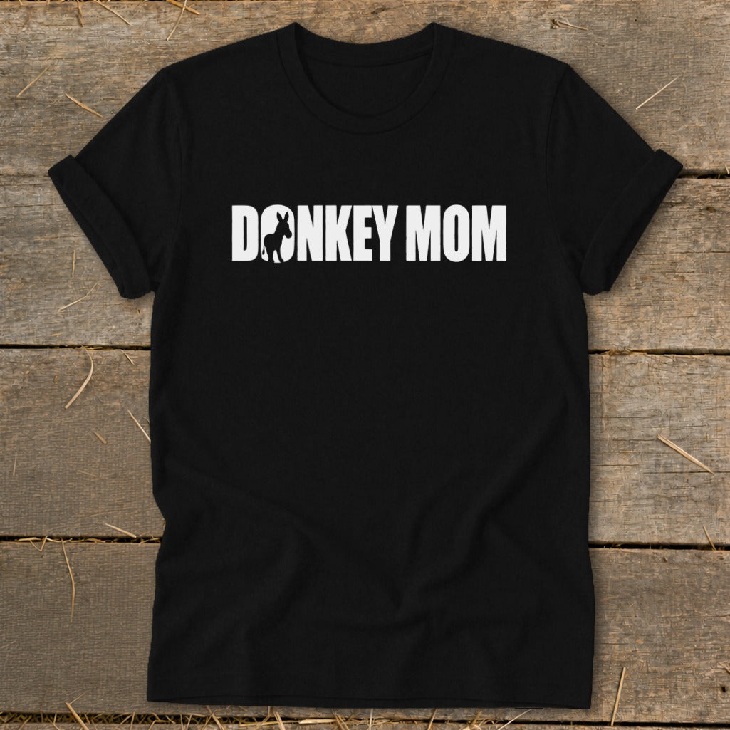 A black t-shirt with rolled-up sleeves, featuring the white text DONKEY MOM, where the O is replaced by a donkey silhouette, resting on a wooden background.