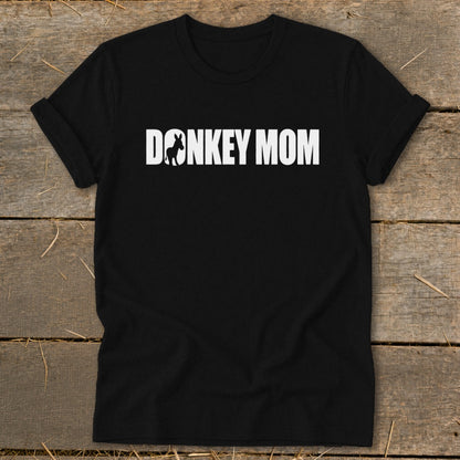 A black t-shirt with rolled-up sleeves, featuring the white text DONKEY MOM, where the O is replaced by a donkey silhouette, resting on a wooden background.