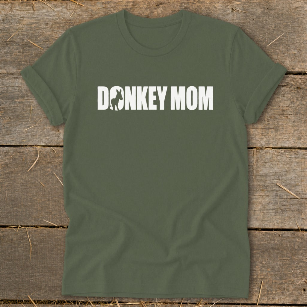 Olive green t-shirt with white text reading "DONKEY MOM" featuring a donkey silhouette inside the letter O, laid flat on a rustic wooden background.