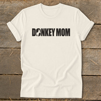A cream-colored t-shirt with rolled-up sleeves lies flat on a rustic wooden surface, featuring black text that reads "DONKEY MOM" with a donkey silhouette inside the first letter O.