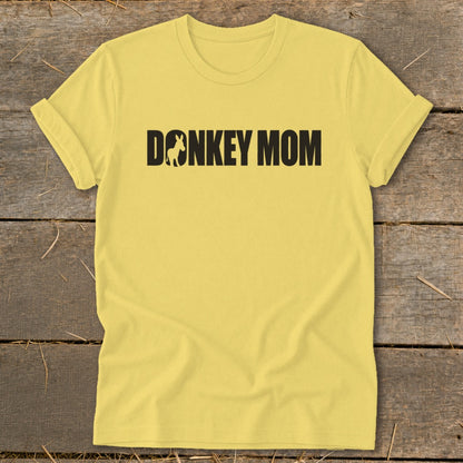 A yellow t-shirt featuring the black text Donkey Mom with a donkey silhouette inside the letter O, laid flat on a rustic wooden background.