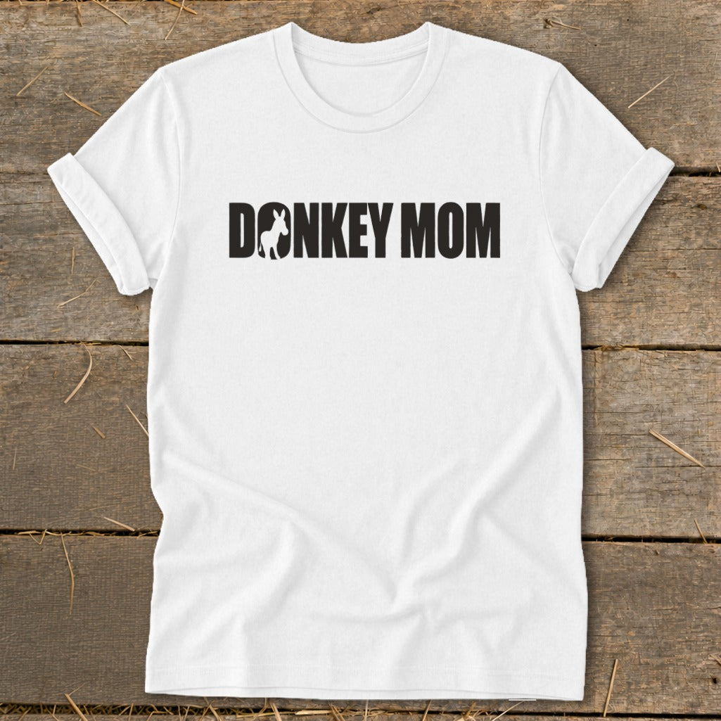 A white crew-neck t-shirt with rolled sleeves lies flat on a wooden surface, featuring black text reading DONKEY MOM with a small donkey silhouette inside the letter O.