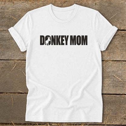 A white crew-neck t-shirt with rolled sleeves lies flat on a wooden surface, featuring black text reading DONKEY MOM with a small donkey silhouette inside the letter O.