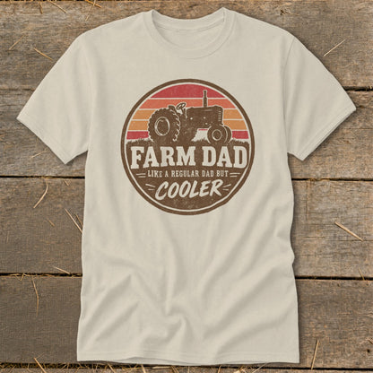 A cream-colored short-sleeved t-shirt features a vintage-style circular graphic of a tractor with sunset stripes and text reading "FARM DAD LIKE A REGULAR DAD BUT COOLER".