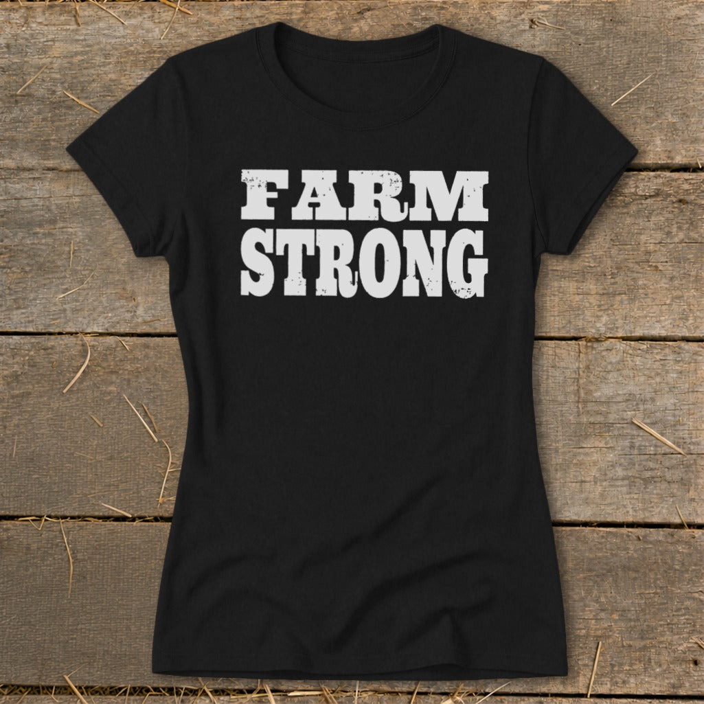 Black short-sleeved t-shirt with distressed white text reading FARM STRONG centered on the chest, laid flat on wooden boards.