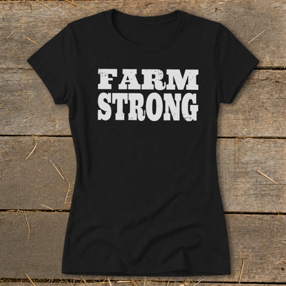 Black short-sleeved t-shirt with distressed white text reading FARM STRONG centered on the chest, laid flat on wooden boards.