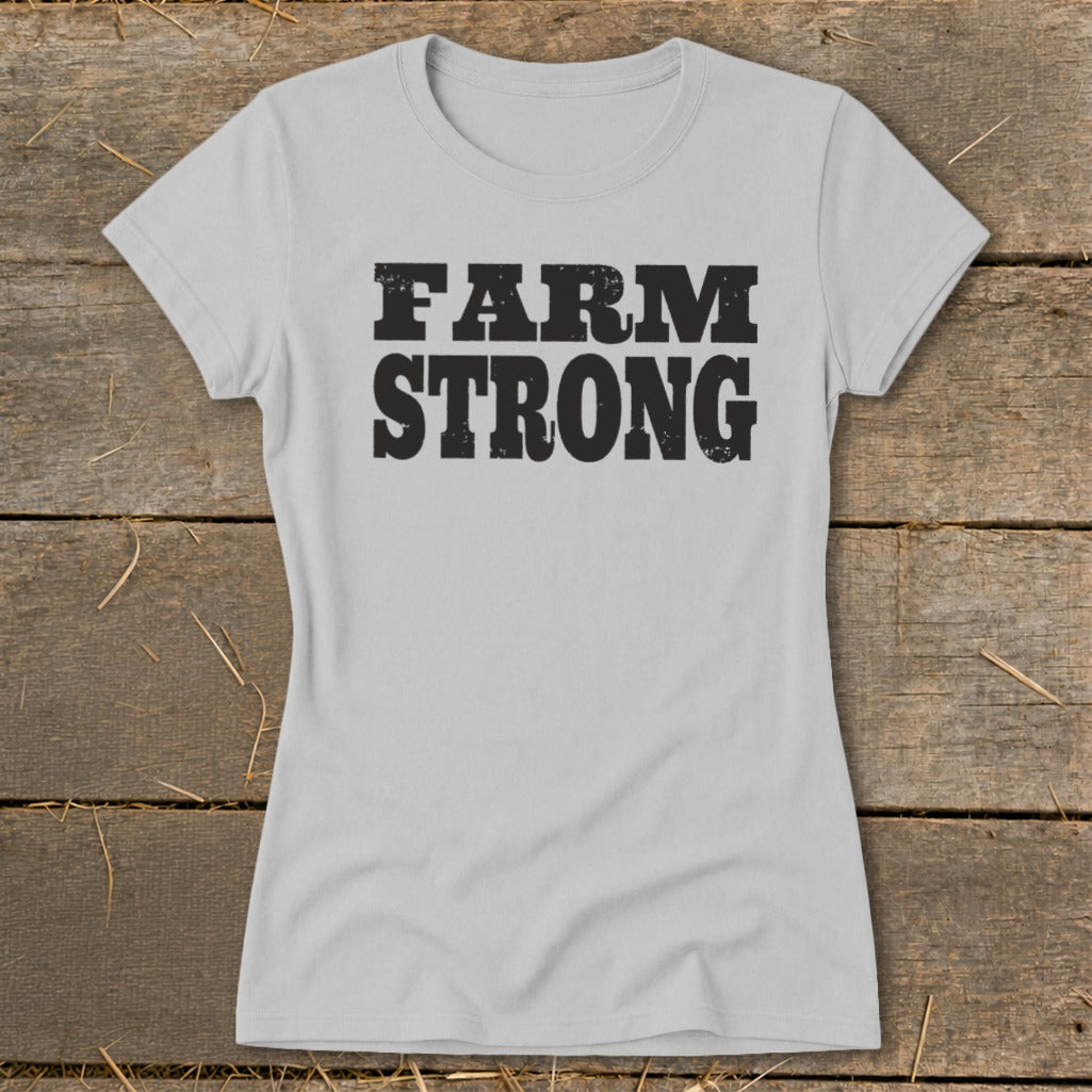 Light grey women's short-sleeve t-shirt with bold black text reading FARM STRONG on a rustic wooden background.