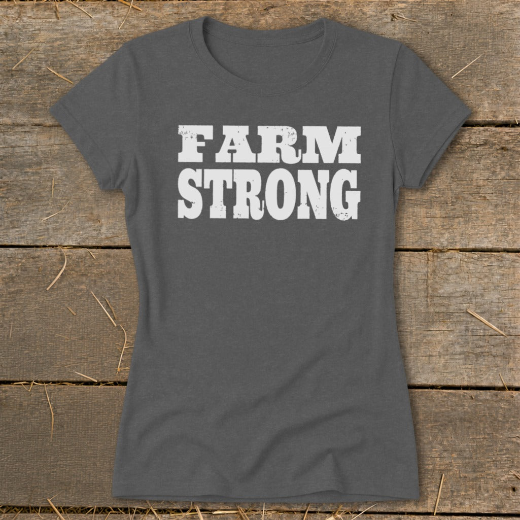 A dark gray women's t-shirt rests on a wooden surface, featuring the white text "FARM STRONG" on the chest.