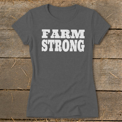 A dark gray women's t-shirt rests on a wooden surface, featuring the white text "FARM STRONG" on the chest.