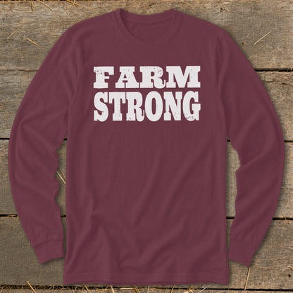 Maroon long-sleeved t-shirt with distressed white text reading FARM STRONG centered on the chest, laid flat on wooden planks.