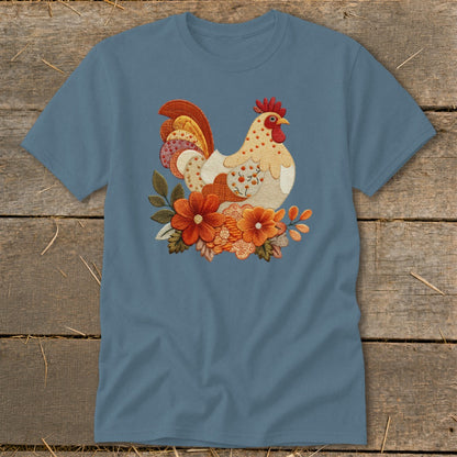 A slate-blue short-sleeve t-shirt features a detailed embroidered rooster graphic surrounded by autumn-colored flowers on a rustic wooden background.