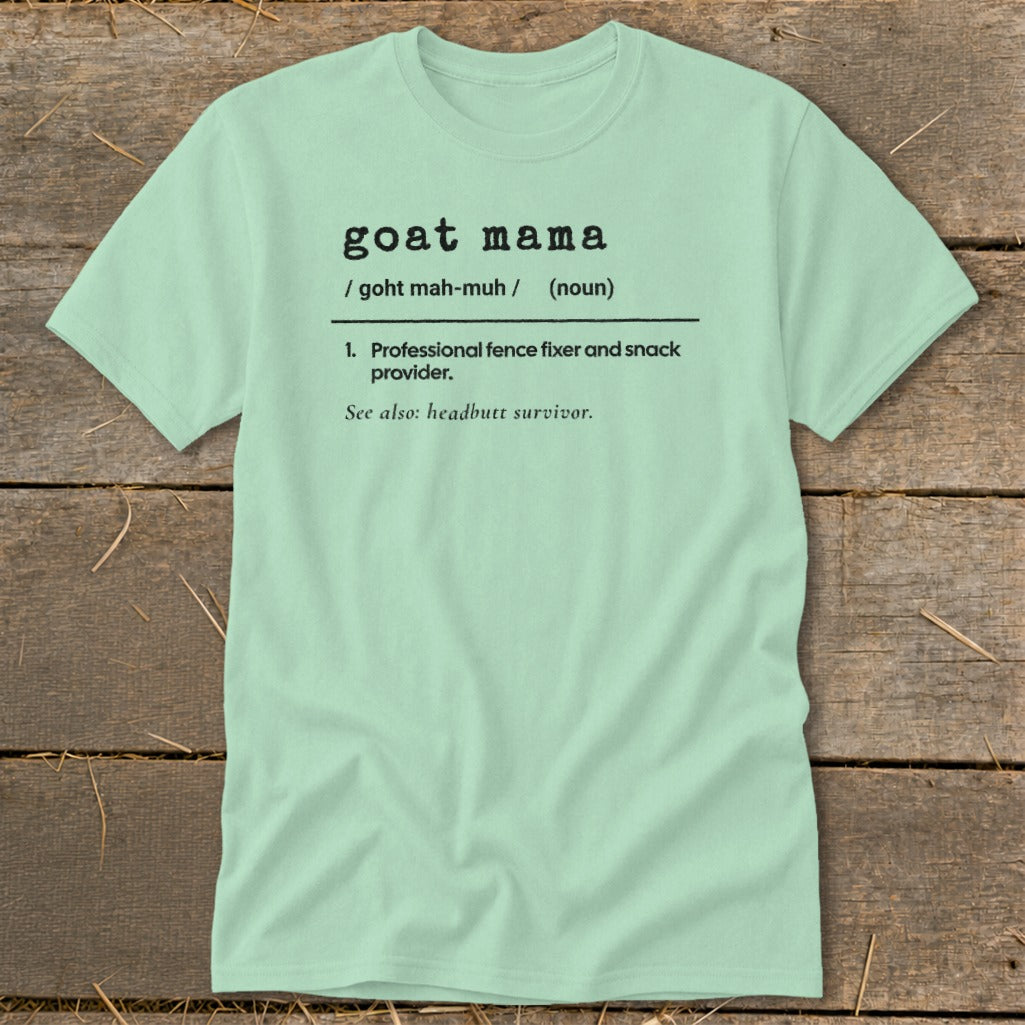 A light green t-shirt with black text that defines "goat mama" laid flat on wooden planks.