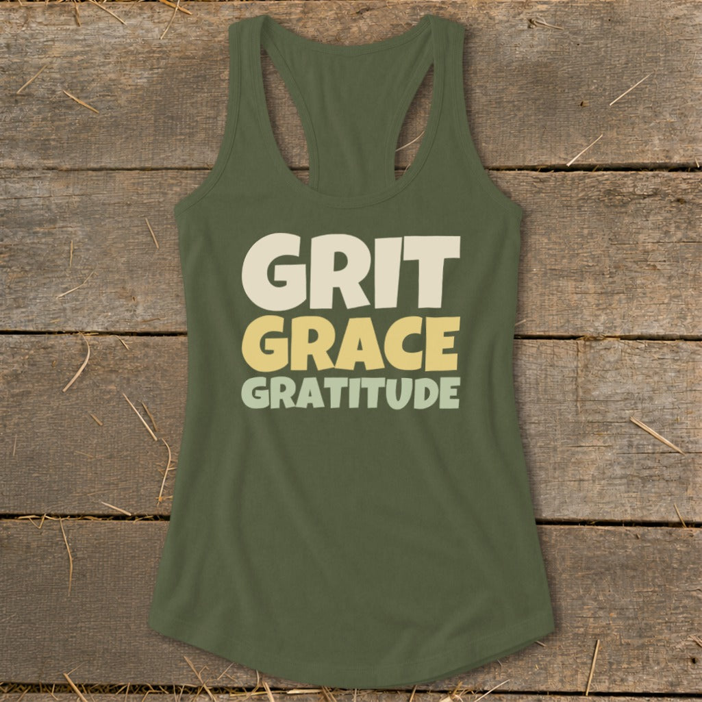 An olive green racerback tank top with the text "GRIT GRACE GRATITUDE" printed across the chest, laid flat on a weathered wooden surface.
