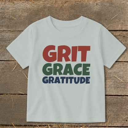 Light gray short-sleeved children's t-shirt printed with the words GRIT GRACE GRATITUDE on a rustic wooden background.