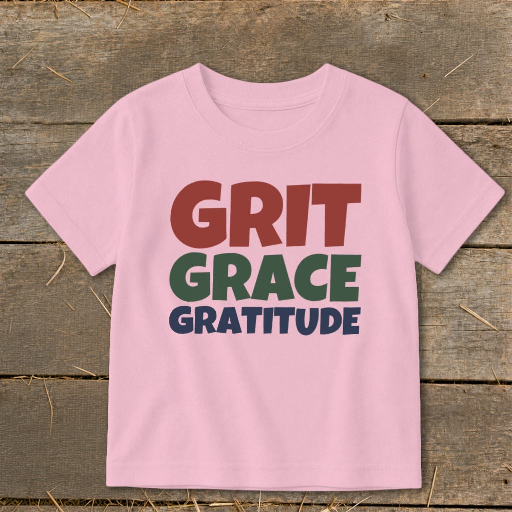 Light pink children's t-shirt printed with the words Grit, Grace, and Gratitude on a rustic wooden background.