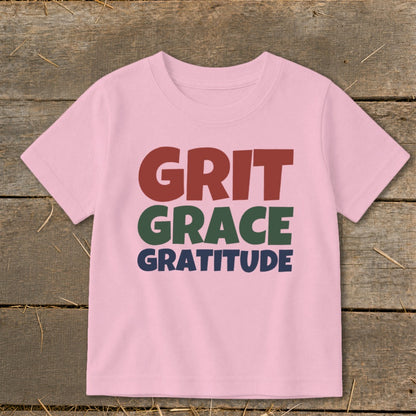Light pink children's t-shirt printed with the words Grit, Grace, and Gratitude on a rustic wooden background.