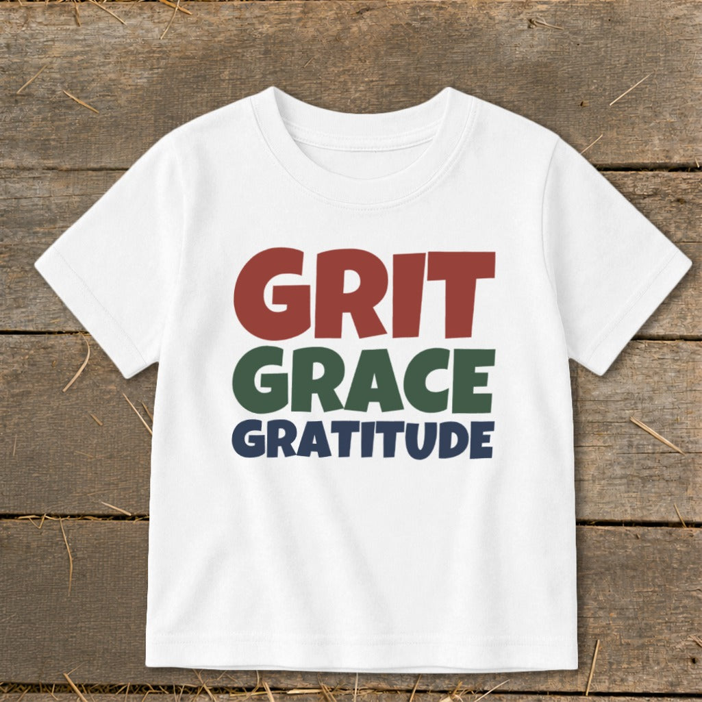 White children's t-shirt printed with the words GRIT in red, GRACE in green, and GRATITUDE in blue.