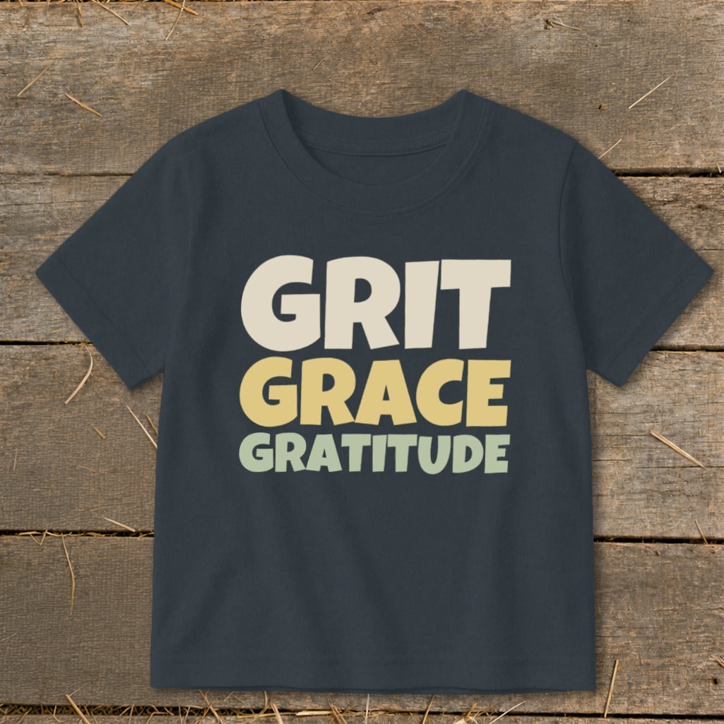A dark gray children's t-shirt printed with the stacked words GRIT, GRACE, and GRATITUDE on a wooden surface.