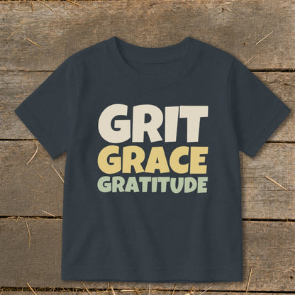 A dark gray children's t-shirt printed with the stacked words GRIT, GRACE, and GRATITUDE on a wooden surface.