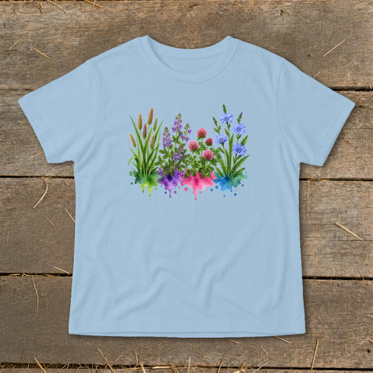 Light blue t-shirt featuring a watercolor botanical illustration of wildflowers with colorful splashes underneath on a wooden background.