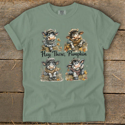 Muted green t-shirt featuring four illustrated cows in autumn hats and scarves with the text Hay There Pumpkin.