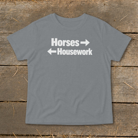 A grey short-sleeved t-shirt with white text reading Horses with a right arrow above Housework with a left arrow, laid flat on a rustic wooden background.