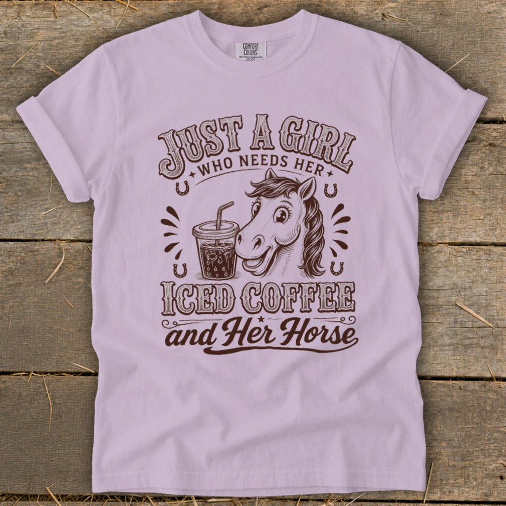 Light purple t-shirt featuring brown text and graphics that read Just a girl who needs her iced coffee and her horse, alongside illustrations of a horse and an iced coffee.