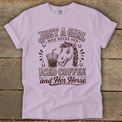 Light purple t-shirt featuring brown text and graphics that read Just a girl who needs her iced coffee and her horse, alongside illustrations of a horse and an iced coffee.