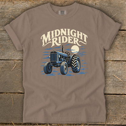 A taupe t-shirt featuring a graphic of a blue tractor under a moon with the text Midnight Rider.