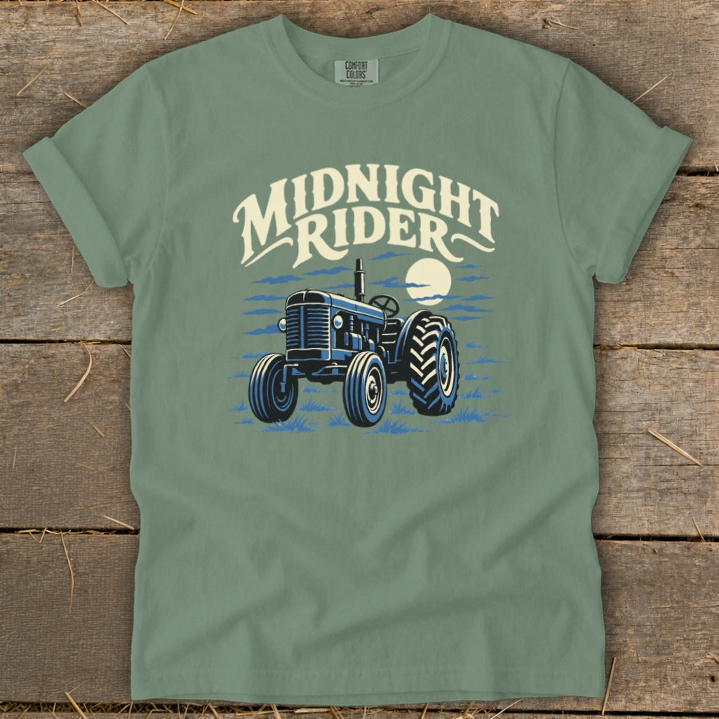 Sage green t-shirt with a blue tractor graphic and the text "Midnight Rider" laid over a wooden surface.