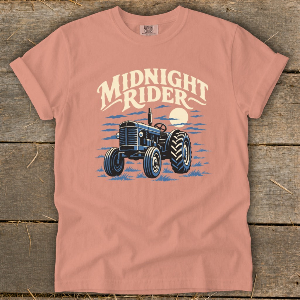 Pink t-shirt with a blue tractor graphic and the text "Midnight Rider" laid flat on wood.