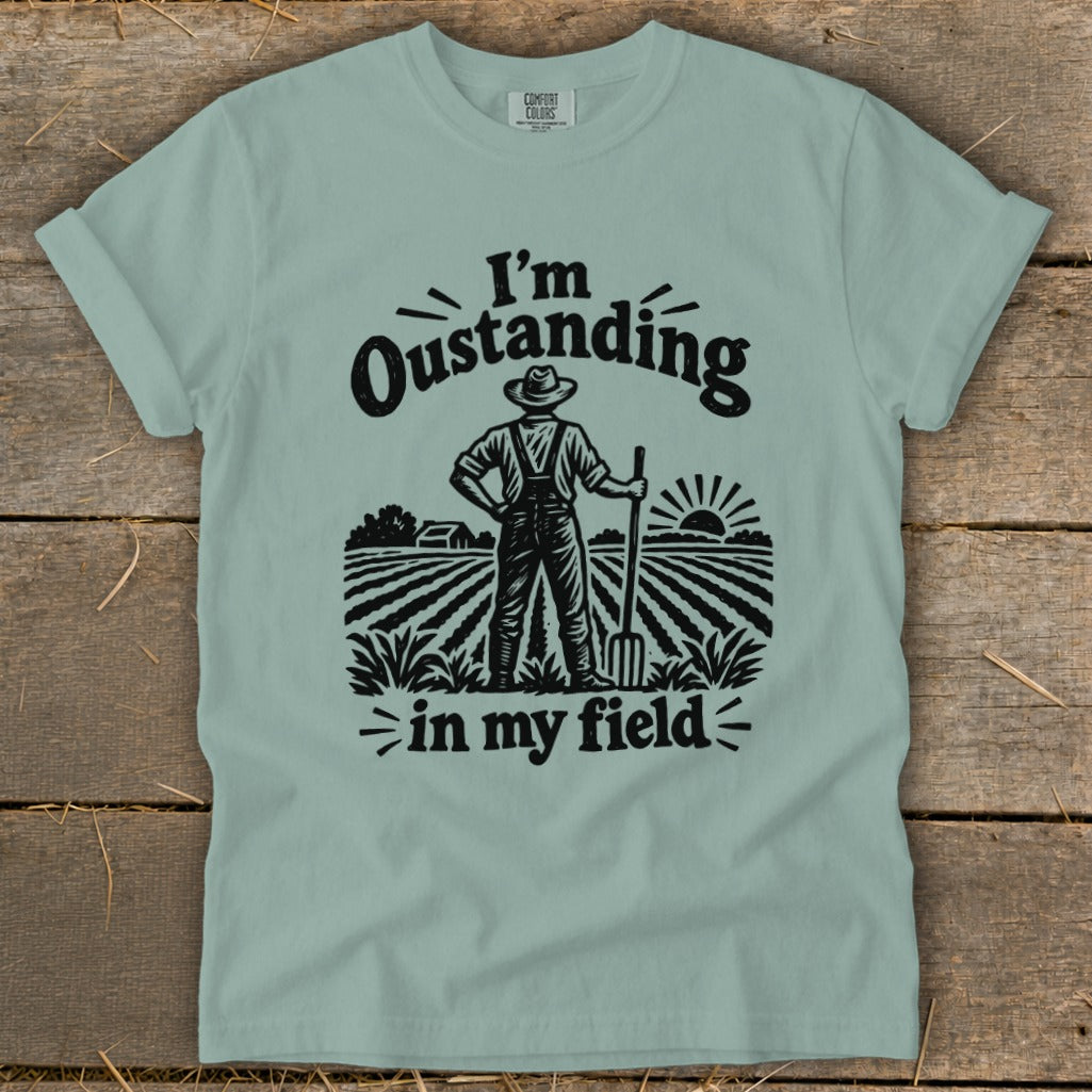 A sage green t-shirt features a black graphic of a farmer holding a pitchfork in a field, with text that reads I'm Oustanding in my field.