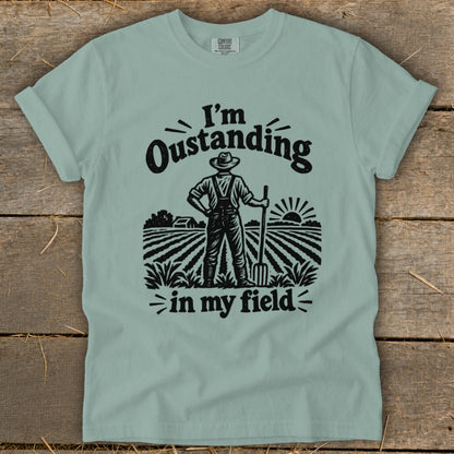 A sage green t-shirt features a black graphic of a farmer holding a pitchfork in a field, with text that reads I'm Oustanding in my field.