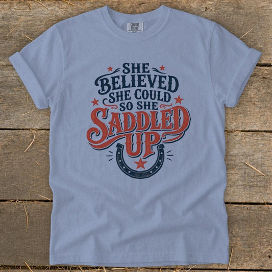 Light blue t-shirt with "She Believed She Could So She Saddled Up" and horseshoe graphic.