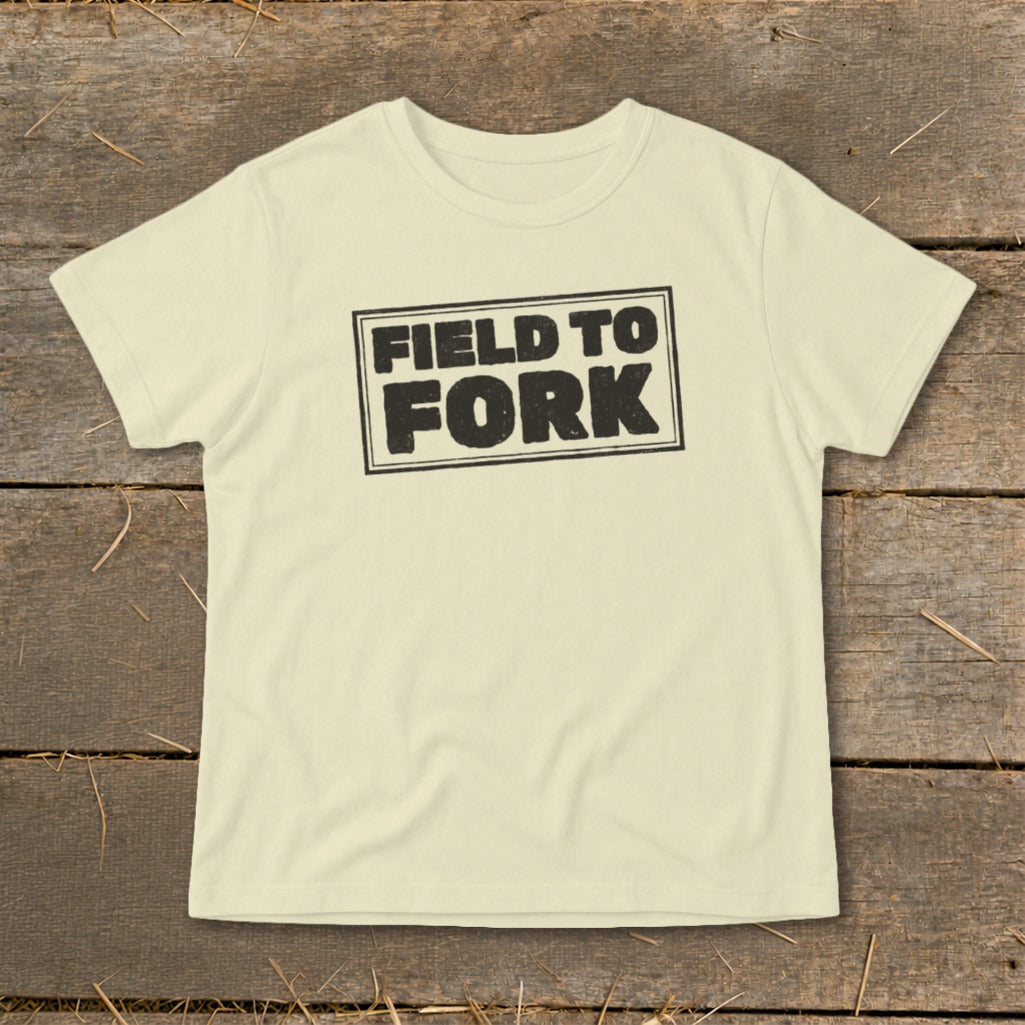 A cream-colored short-sleeved t-shirt featuring the black text FIELD TO FORK laid flat on a rustic wooden background.