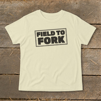 A cream-colored short-sleeved t-shirt featuring the black text FIELD TO FORK laid flat on a rustic wooden background.