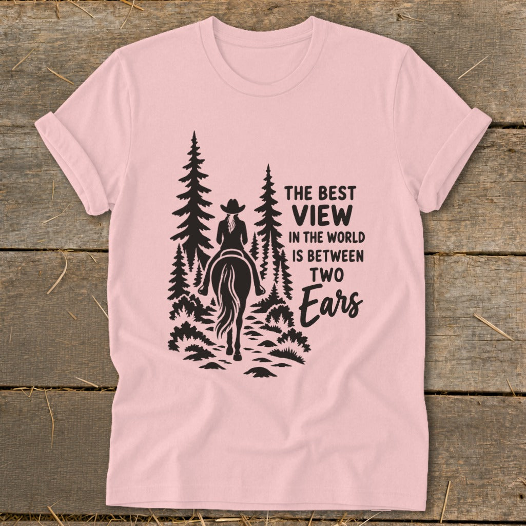 A light pink t-shirt featuring a black graphic of a horse rider in the forest with the text The Best View in the World is Between Two Ears.