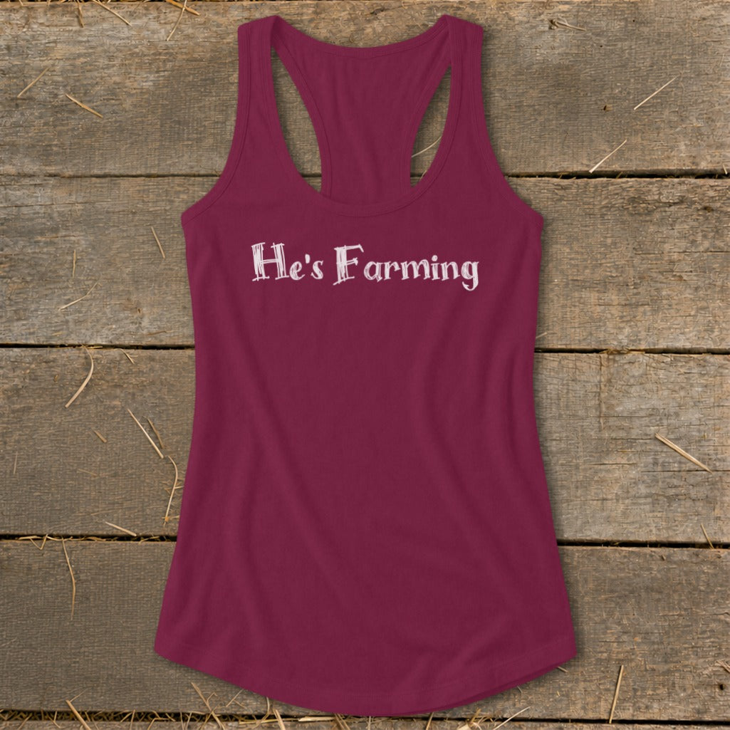 Maroon tank top with white text that reads "He's Farming" on a wooden background.