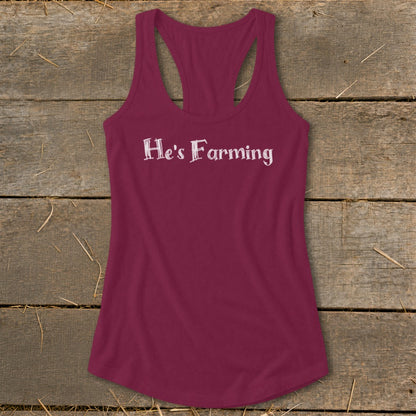 Maroon tank top with white text that reads "He's Farming" on a wooden background.